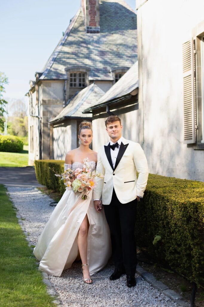 Castle Wedding Venues in New England | Kelsey Sheehan Photography Rhode Island Wedding Photographer | A couple stands elegantly in front of a stone building with greenery. The woman in a strapless gown holds a bouquet, and the man wears a white tuxedo.