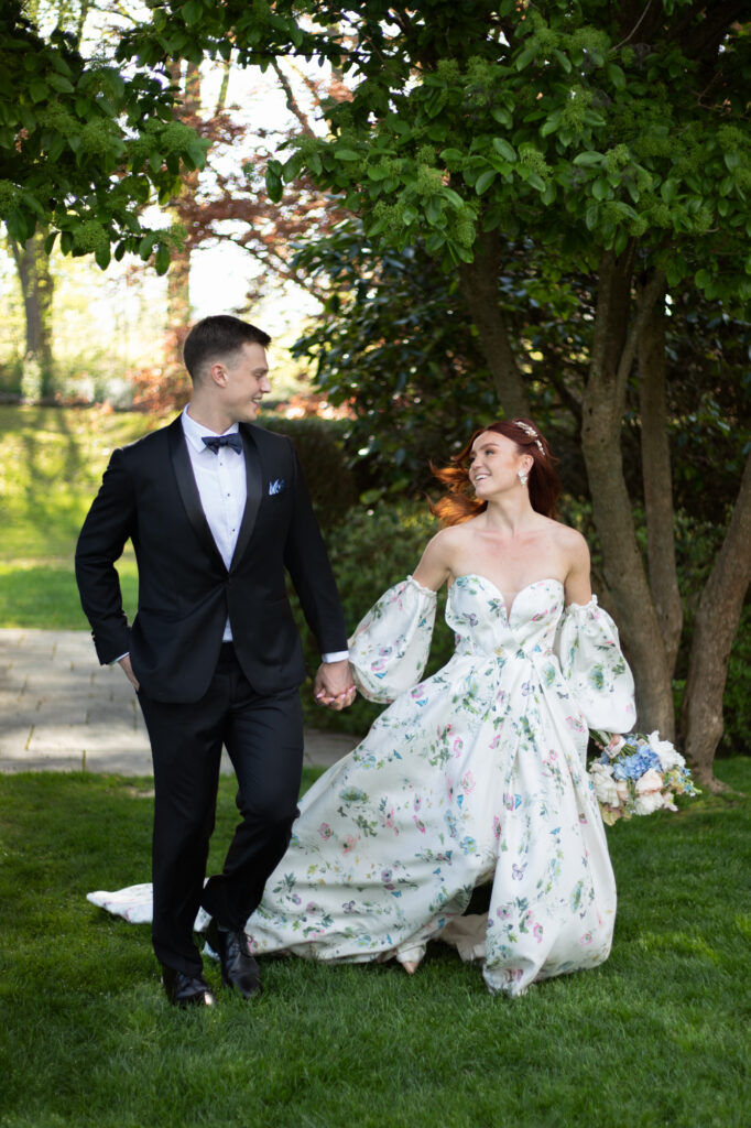 Glen Manor House | Kelsey Sheehan Photography Rhode Island Wedding Photographer | A joyful couple holds hands, walking through a sunlit garden. The bride wears a floral gown, while the groom is in a black tuxedo. They share a happy glance.