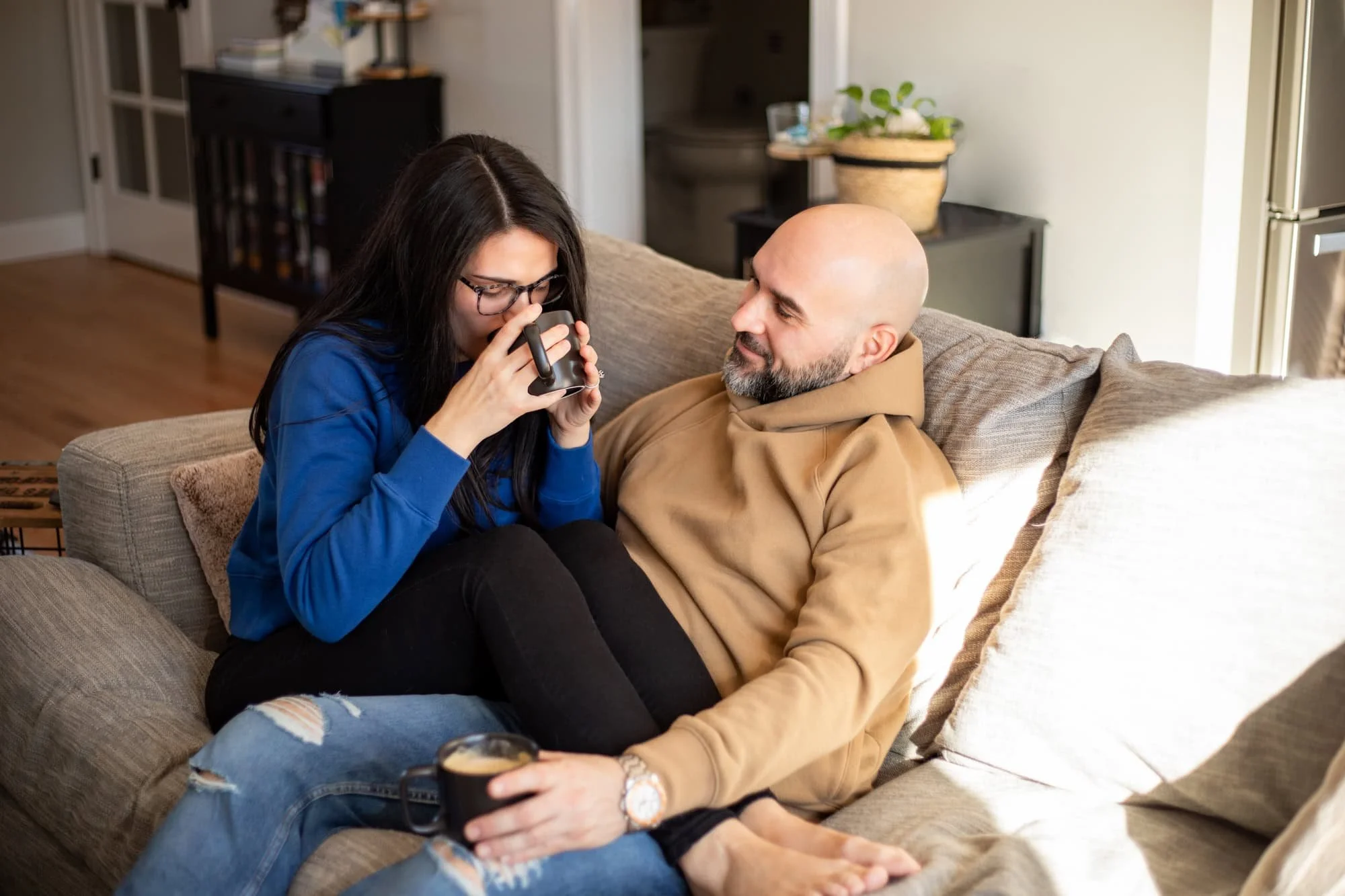 A couple sitting on a couch, one person with a bald head wearing a brown hoodie and the other in a blue sweatshirt and glasses, both holding black mugs with beverages. The room has neutral-toned walls and a small plant on a nearby table.