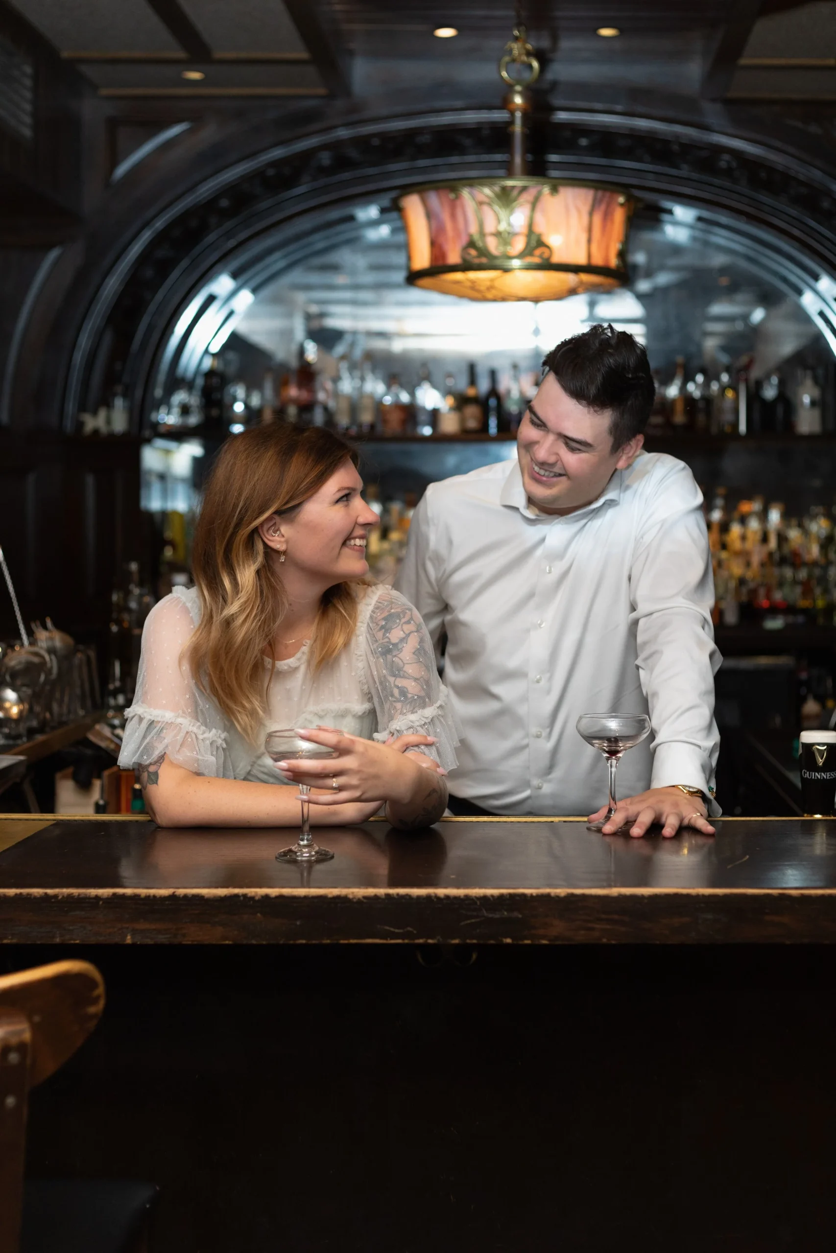 Couple looking at each other while standing behind the bar in Boston, MA after they just got engaged. She is wearing a vintage lace off white dress. He is wearing a white button up shirt. 