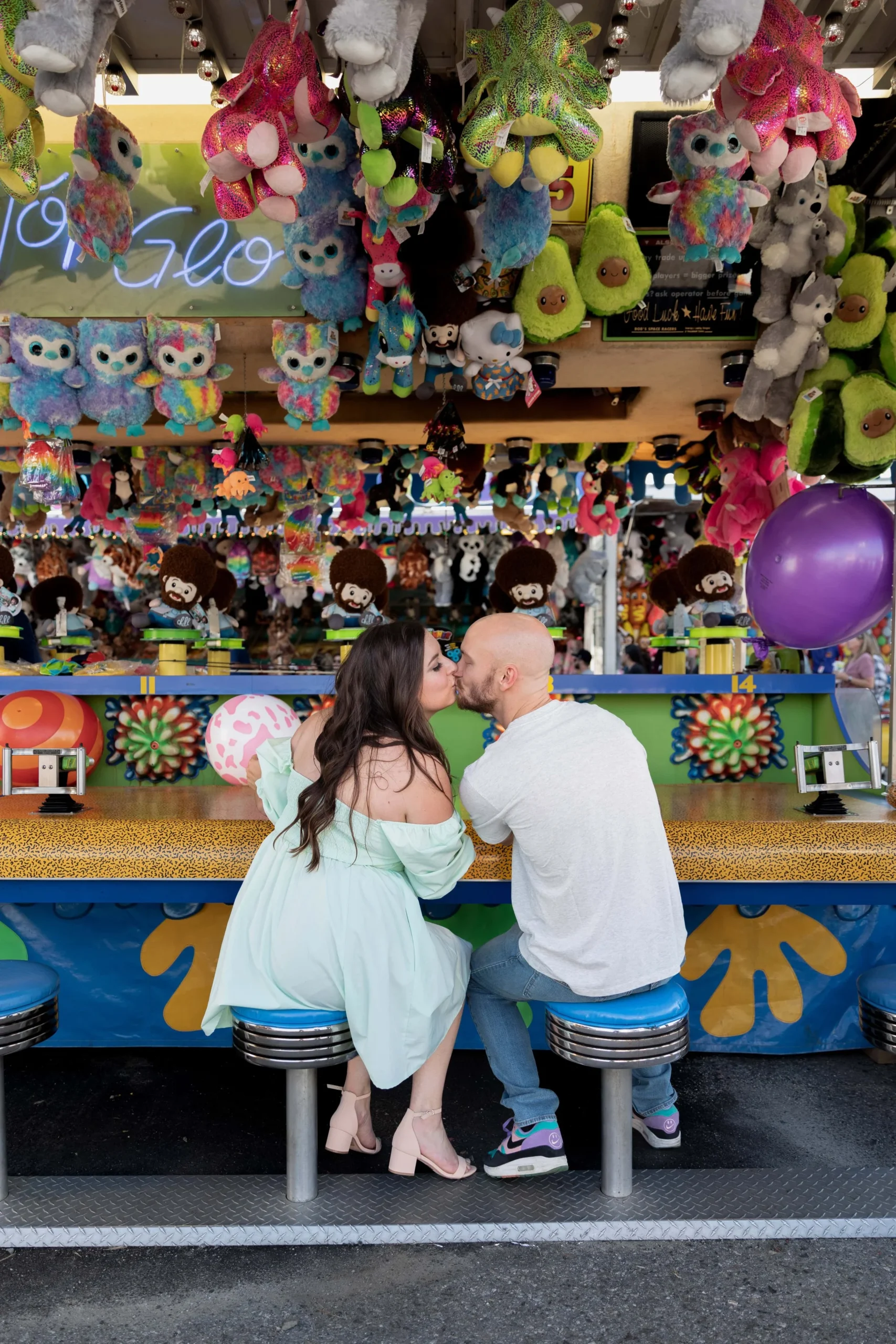 Couple kissing in front of the water blast game at the Topsfield Fair in Massachusetts. She is wearing a light teal off the shoulder dress and he is wearing jeans and a white polo shirt. 