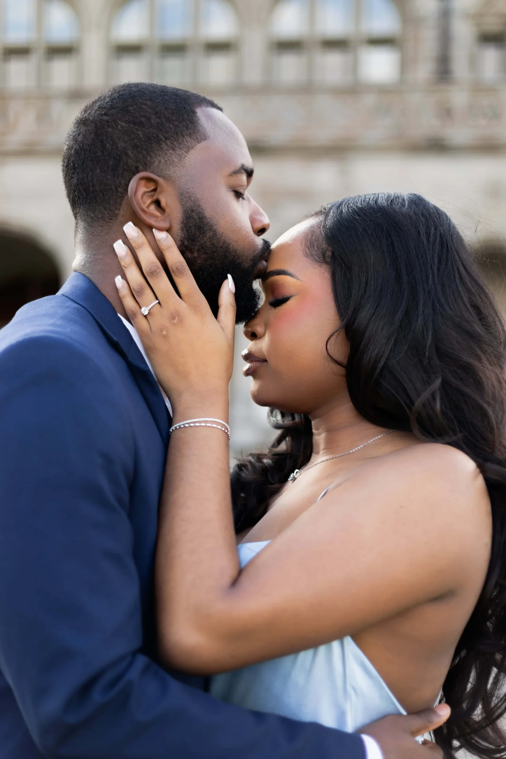 Guy is kissing his fiance on the forehead behind a mansion in Newport, Rhode Island. He is wearing a dark blue suit and she is wearing a light blue satin dress. 