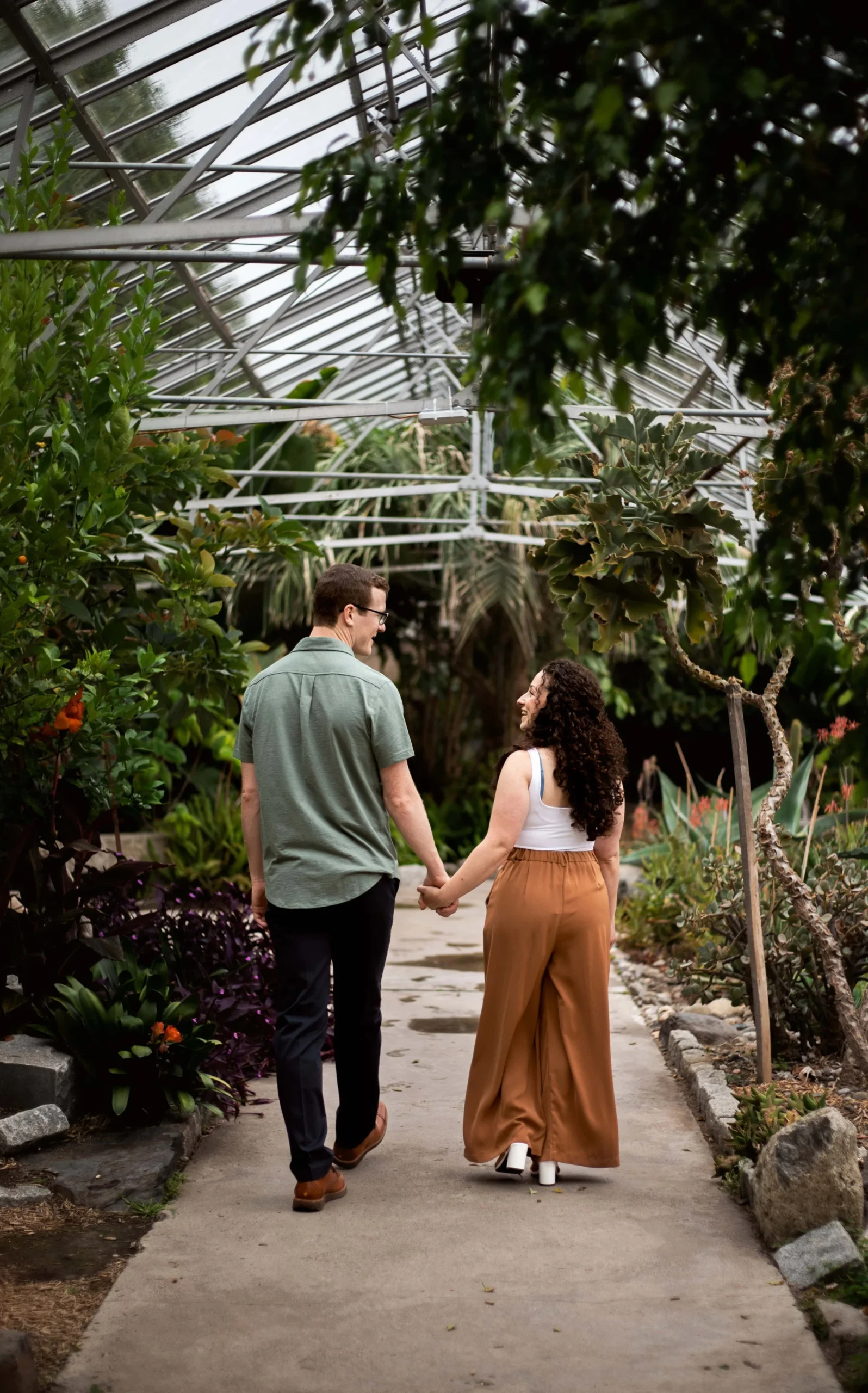 Couple walking hand in hand at a botanical garden in Rhode Island. She is wearing a white tank top and a wide leg orange pant. He is wearing a sage green top and black pants. 