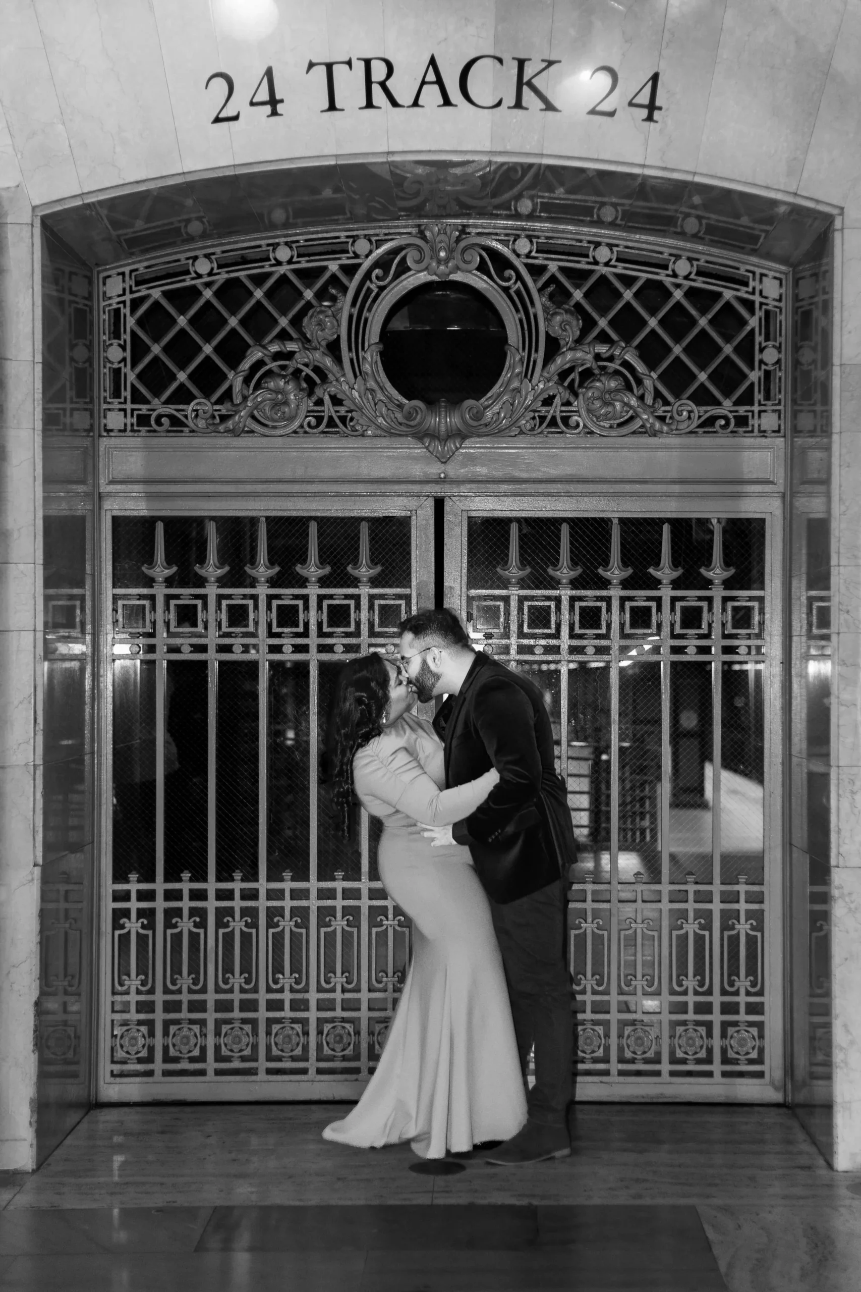 Black and white image of a couple kissing in front of track 24 at Grand Central Station in New York City. He is wearing a suit and she is wearing a bright red fitted dress. 
