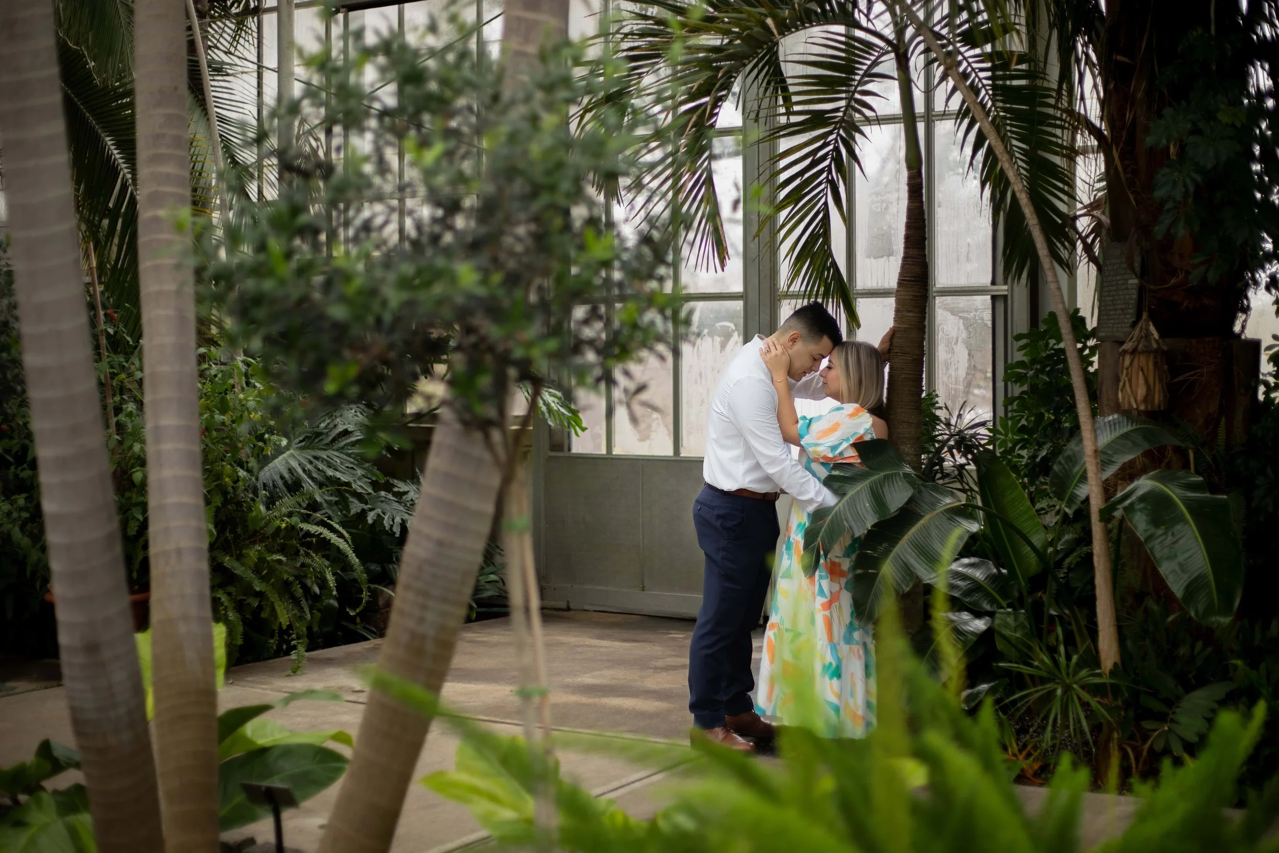 Couple leaning against a tree at the Roger Williams Botanical Gardens. He is wearing navy pants and a long sleeve white dress shirt. She is wearing a flowy dress that features teal and orange pattern. 