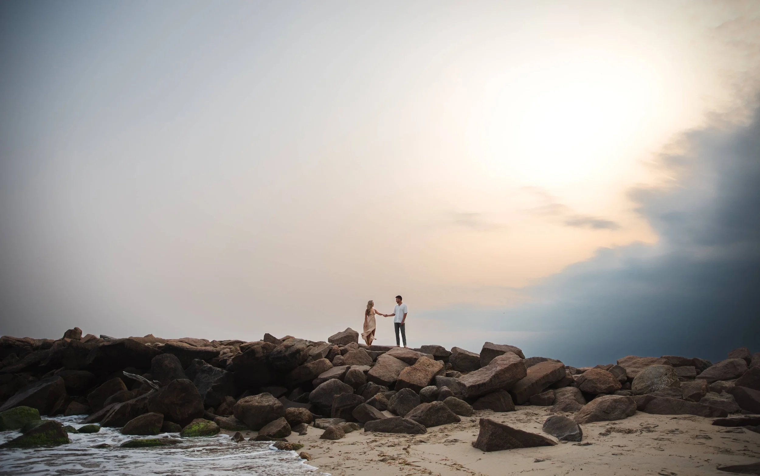 Landsape photo of a couple walking along a jetty in Rhode Island. The sun is starting to set in the thunderstorm clouds. She is wearing a peach dress and he is wearing dress pants and a button up shirt. 