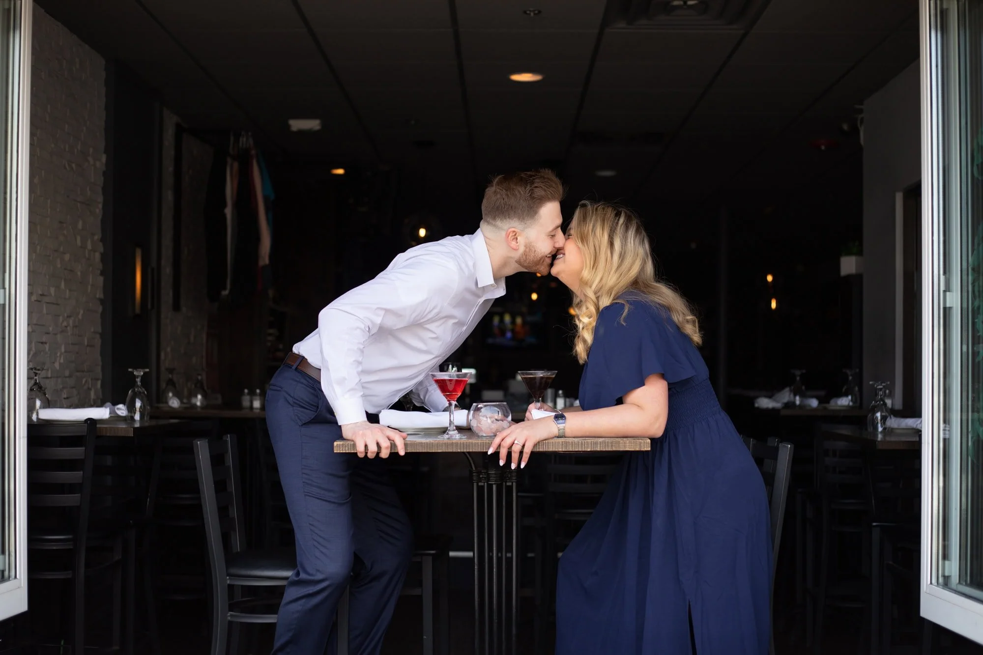 Couple sitting at a restaurant in Plymouth, MA drinking martinis. Girl is wearing a long dark blue dress. The guy is wearing navy blue pants and a white button up shirt. He is leaning across the table and giving her a kiss. 