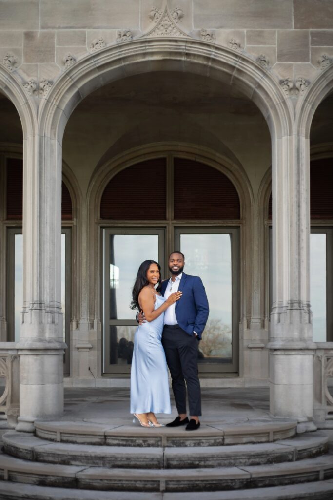 Ochre Court Newport | Kelsey Sheehan Photography Rhode Island Wedding Photographer | A couple stands on elegant stone steps under an ornate archway. She wears a light blue dress, and he dons a blue suit, both smiling warmly. Romantic and sophisticated atmosphere.