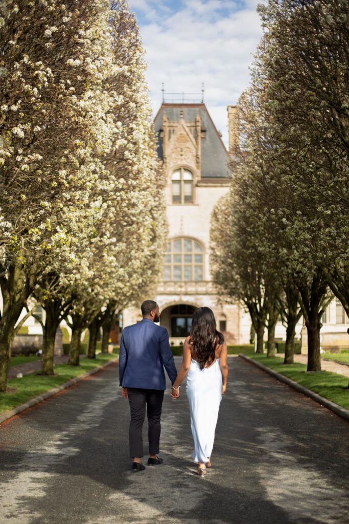 Ochre Court Newport | Kelsey Sheehan Photography Rhode Island Wedding Photographer | A couple walks hand-in-hand down a tree-lined path towards a grand, historic building. The scene conveys romance and tranquility under a clear sky.