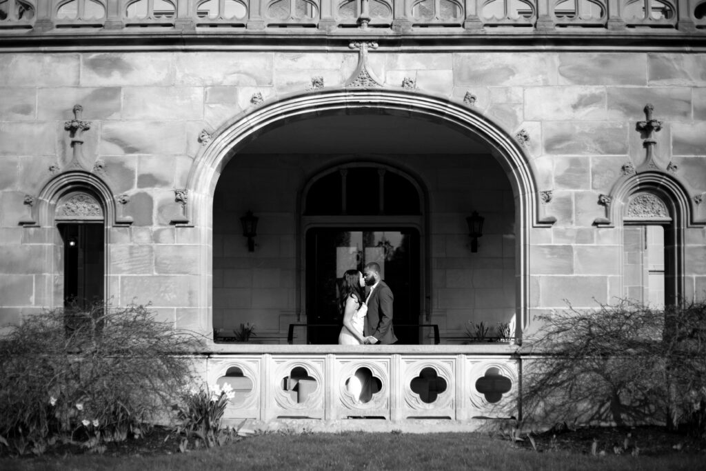 Ochre Court Newport | Kelsey Sheehan Photography Rhode Island Wedding Photographer | Black and white photo of a couple embracing under an ornate stone archway. They stand on a veranda, framed by intricate masonry, creating a romantic setting.
