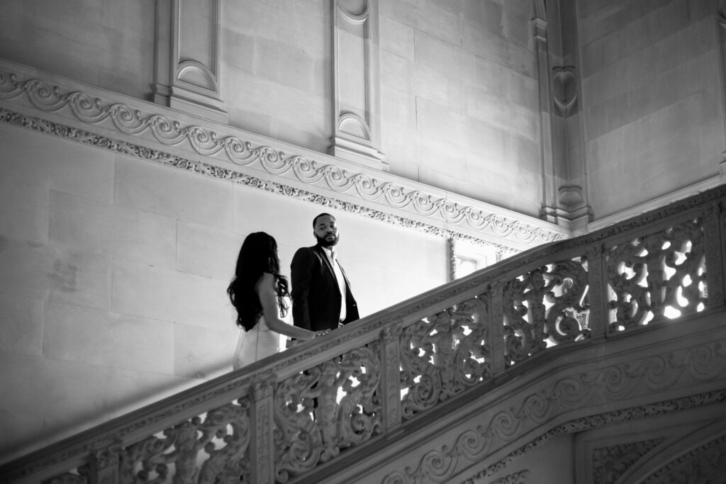 Ochre Court Newport | Kelsey Sheehan Photography Rhode Island Wedding Photographer | A couple ascends an ornate staircase in a grand hall, the woman in a dress and the man in a suit, conveying elegance and romance. Black and white.