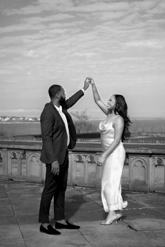 Ochre Court Newport | Kelsey Sheehan Photography Rhode Island Wedding Photographer | A couple dances joyfully on a terrace under a cloudy sky. The man in a suit holds the smiling woman's hand, who wears a flowing dress. They exude happiness and connection.