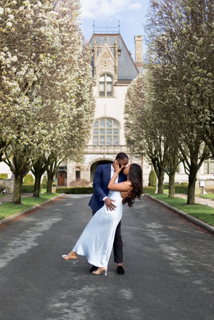 Ochre Court Newport | Kelsey Sheehan Photography Rhode Island Wedding Photographer | A couple shares a romantic embrace on a tree-lined path in front of a grand historic building. The scene conveys elegance and love.