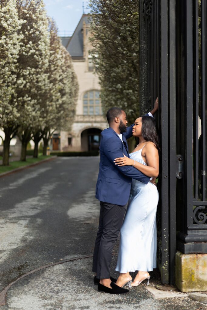 Ochre Court Newport | Kelsey Sheehan Photography Rhode Island Wedding Photographer | A couple embraces near an ornate gate with blooming trees lining a pathway. The man is in a blue suit, and the woman wears a light blue dress, conveying romance.