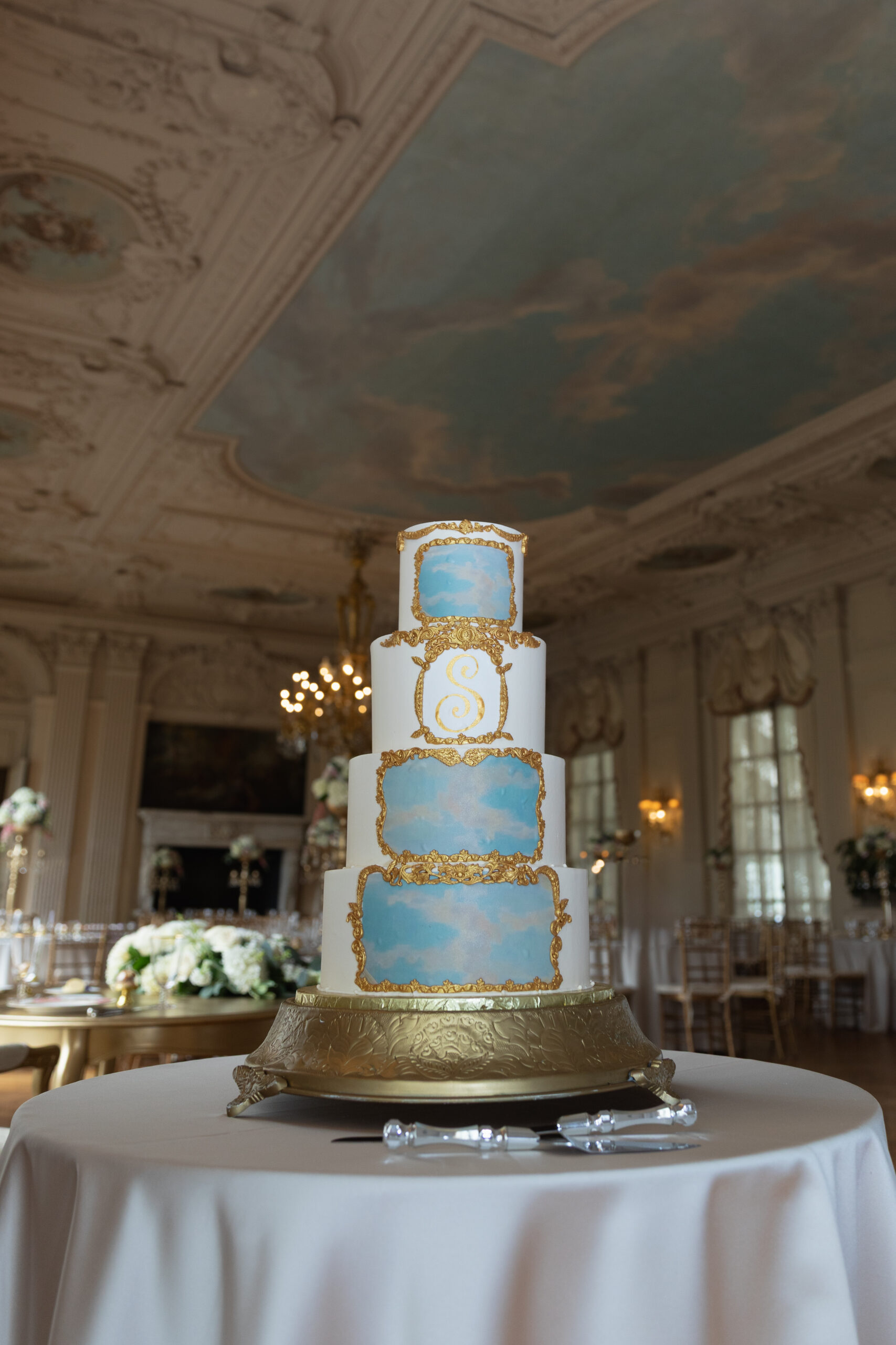 Rosecliff Mansion | Kelsey Sheehan Photography Rhode Island Wedding Photographer | Elegant four-tiered wedding cake with blue and gold ornate designs on a gold stand in a lavish ballroom setting with chandeliers and floral decor.