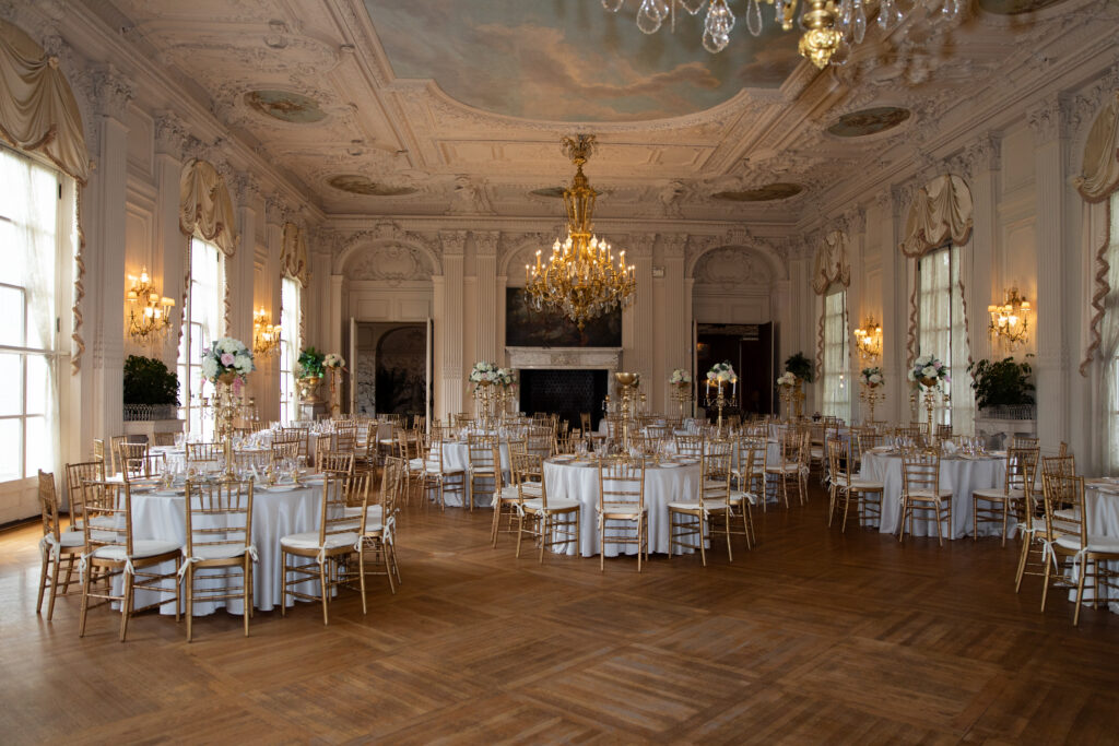 Castle Wedding Venues in New England | Kelsey Sheehan Photography Rhode Island Wedding Photographer | Elegant ballroom with ornate ceiling and large chandelier. Round tables with white tablecloths and gold chairs create a luxurious ambiance.