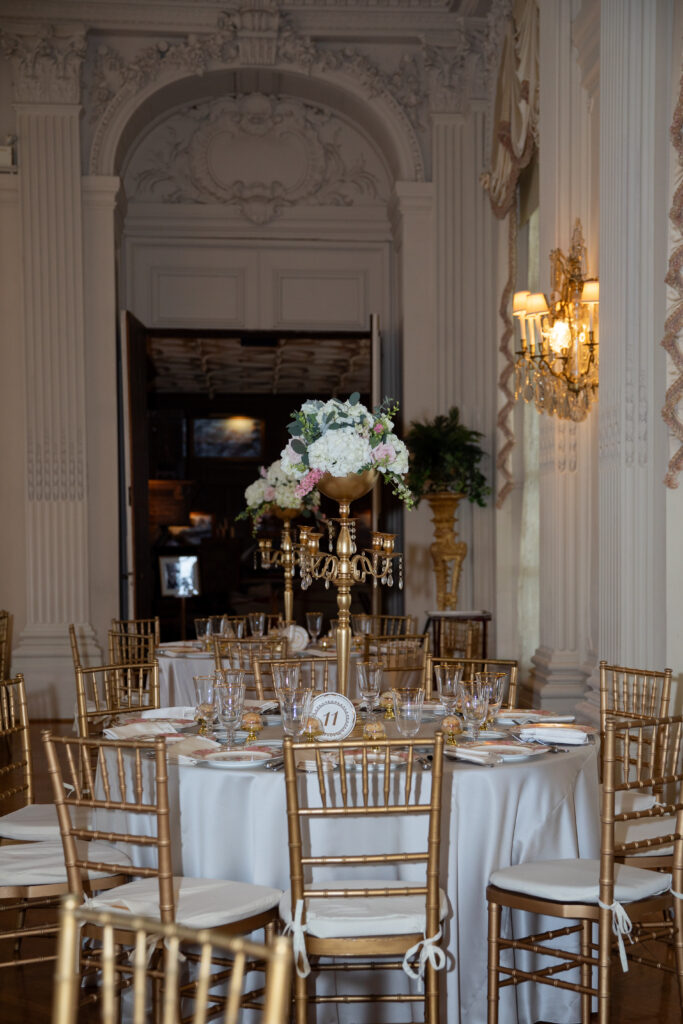 Rosecliff Mansion | Kelsey Sheehan Photography Rhode Island Wedding Photographer | Elegant dining room with a round table set for a formal event. Gold chairs surround a white tablecloth adorned with glassware and a tall floral centerpiece under ornate lighting.