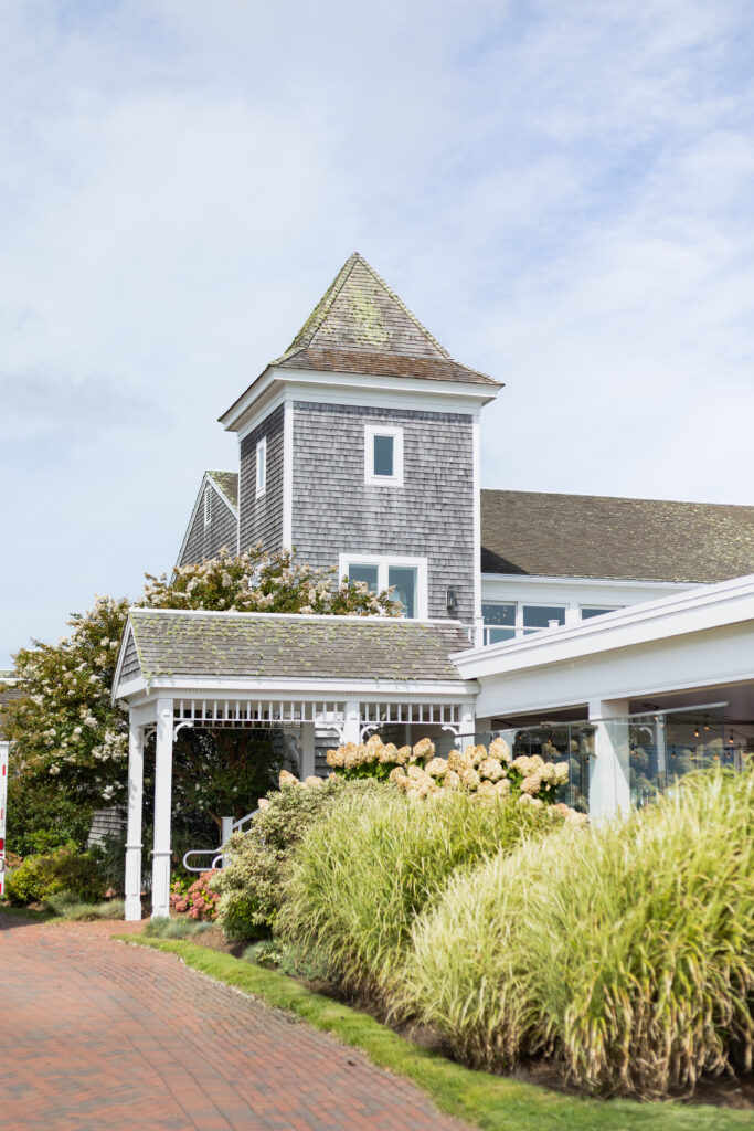 Wychmere Beach Club | Kelsey Sheehan Photography Rhode Island Wedding Photographer | A charming grey shingle house with a pointed roof and white trim sits amid lush gardens with blooming flowers and tall grasses, under a cloudy sky.