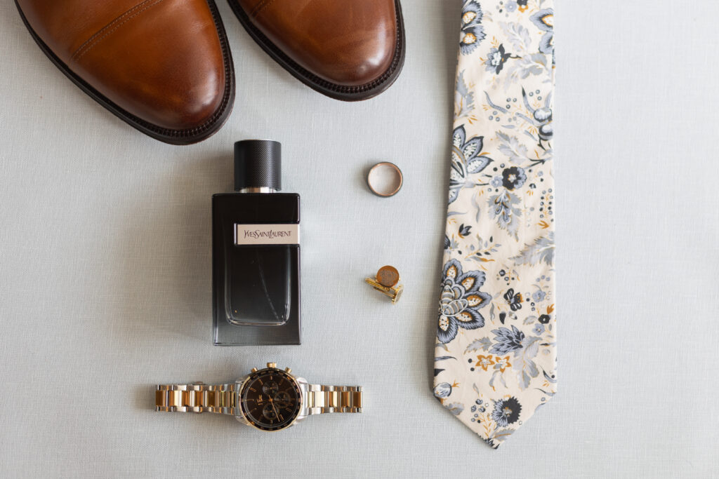 Wychmere Beach Club | Kelsey Sheehan Photography Rhode Island Wedding Photographer | Brown leather shoes, floral tie, black cologne bottle, ring, cufflinks, and a gold watch on a light background. Elegant and sophisticated.