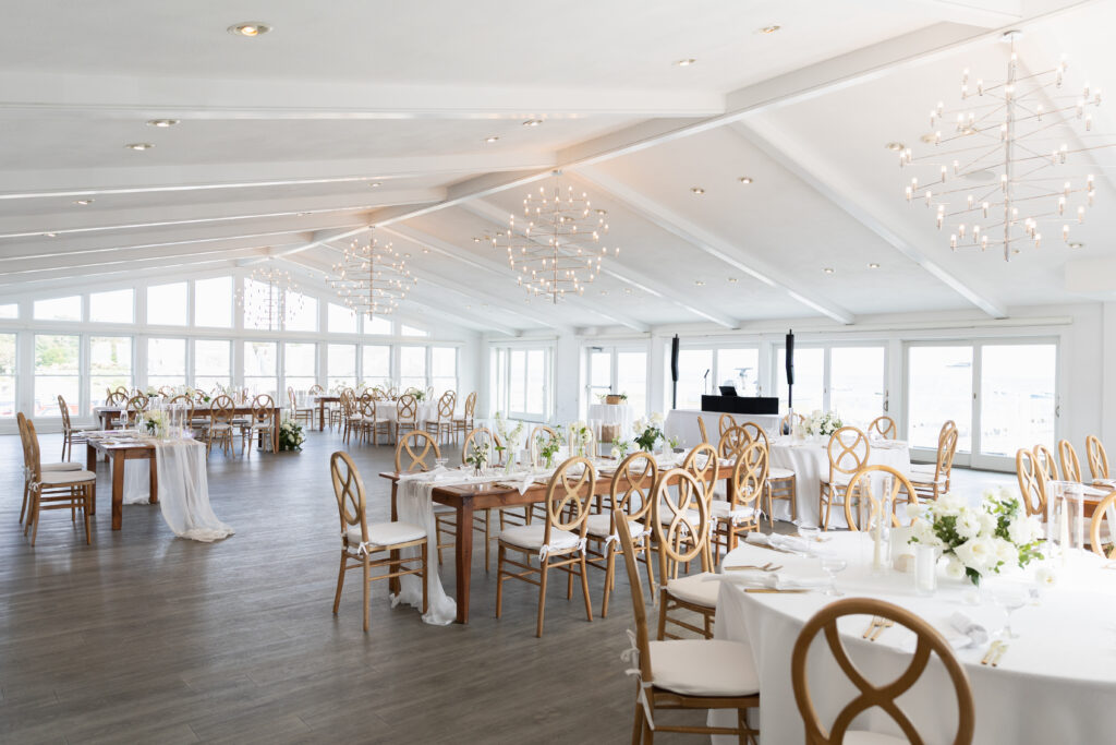 Wychmere Beach Club | Kelsey Sheehan Photography Rhode Island Wedding Photographer | Elegant banquet hall with white walls and wooden floors features round tables with white tablecloths, wooden chairs, floral centerpieces, and modern chandeliers.