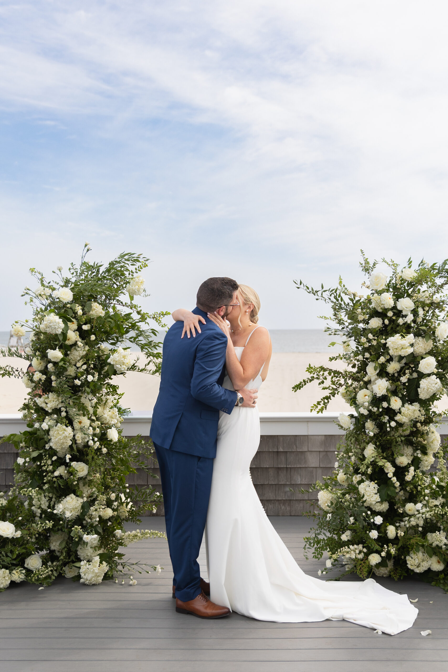 Wychmere Beach Club | Kelsey Sheehan Photography Rhode Island Wedding Photographer | A couple kisses during a seaside wedding, surrounded by lush white floral arrangements. The sky is partly cloudy, adding a serene backdrop to the scene.