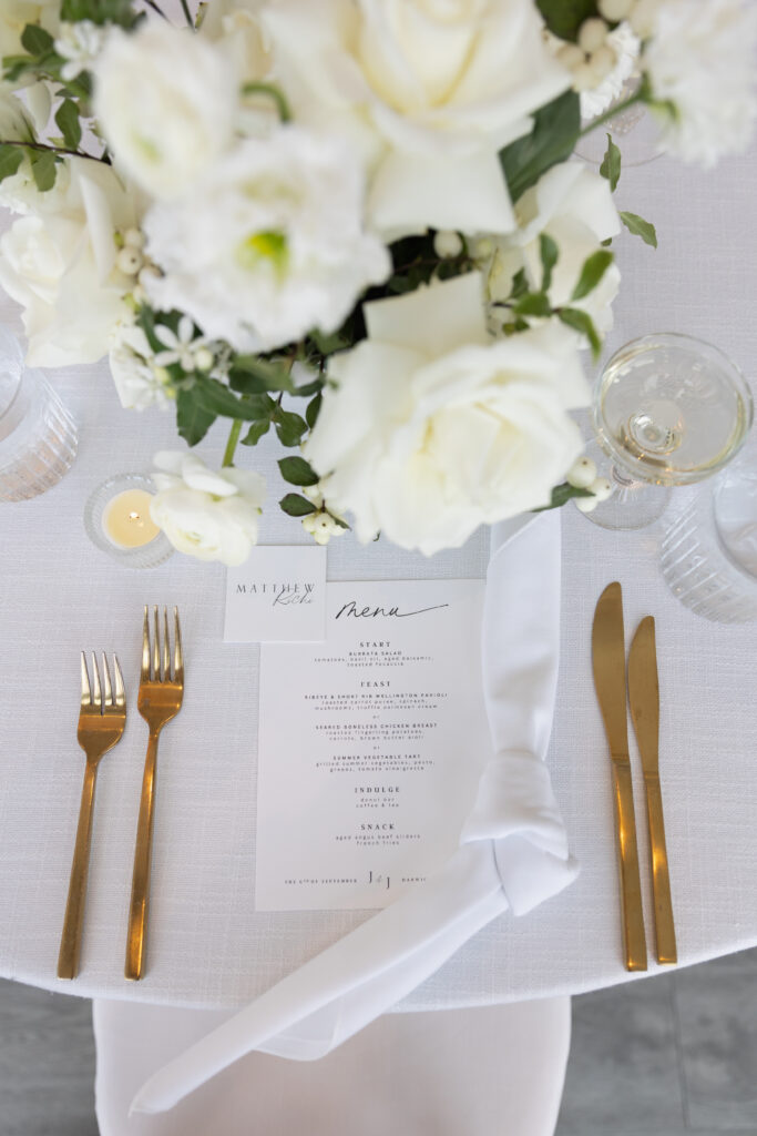 Wychmere Beach Club | Kelsey Sheehan Photography Rhode Island Wedding Photographer | Elegant table setting with white roses centerpiece, white tablecloth, gold cutlery, and a neatly tied napkin. Menu card placed under soft candlelight.