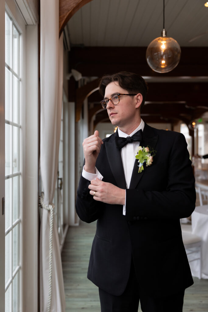 Wychmere Beach Club | Kelsey Sheehan Photography Rhode Island Wedding Photographer | Man in a black tuxedo with a floral boutonniere gazes out a window, adjusting his cufflink. The setting is a refined hall with soft lighting.