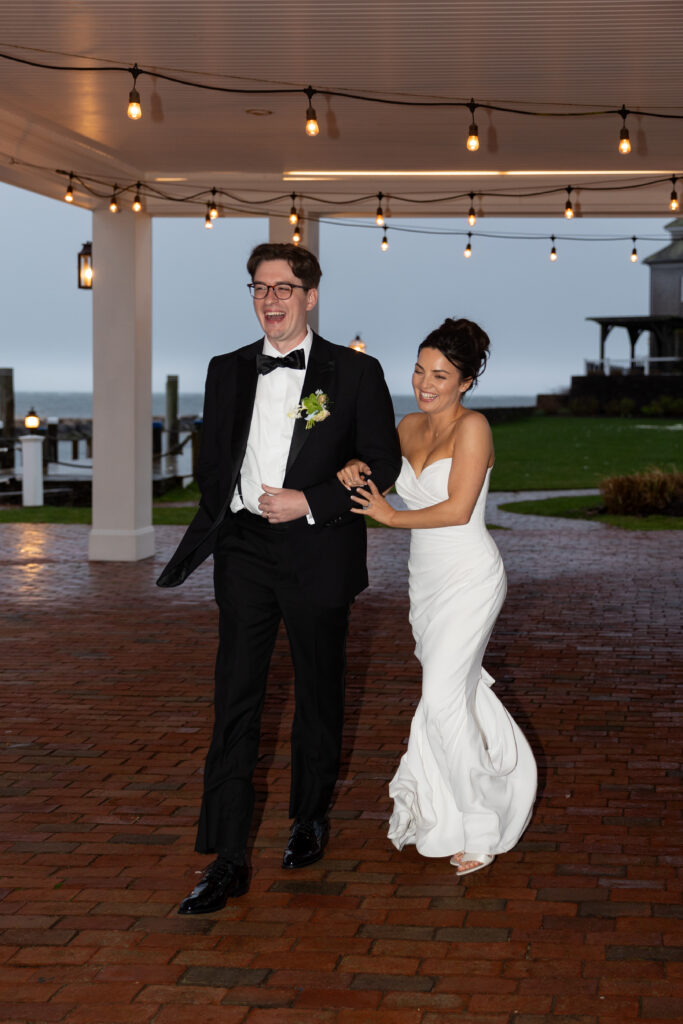 Wychmere Beach Club | Kelsey Sheehan Photography Rhode Island Wedding Photographer | A joyful couple in formal wedding attire laughs together under string lights on a brick patio. The background shows a seaside view and a lawn.