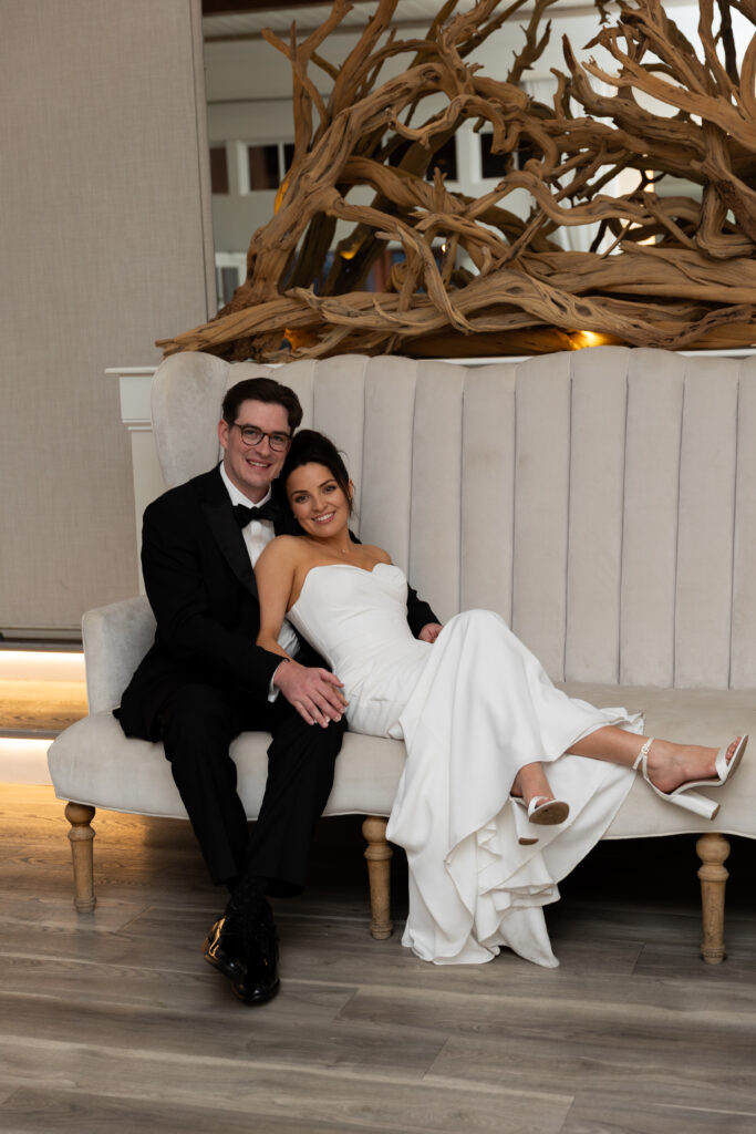 Wychmere Beach Club | Kelsey Sheehan Photography Rhode Island Wedding Photographer | A smiling couple sits on a plush, light-colored bench with driftwood decor behind. The man in a tuxedo embraces the woman in a white dress, exuding joy.