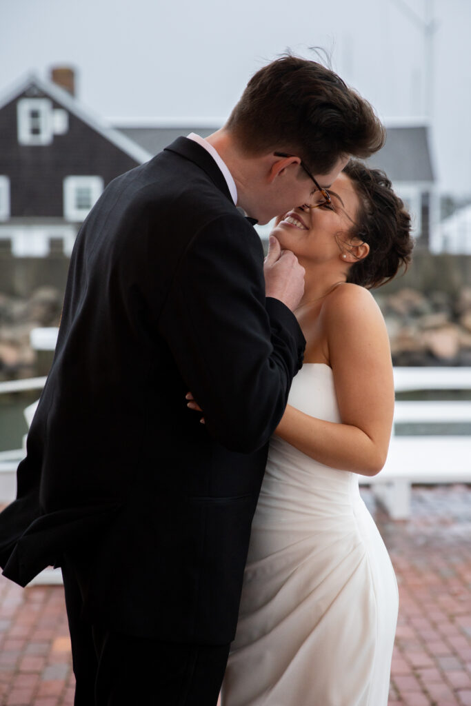 Wychmere Beach Club | Kelsey Sheehan Photography Rhode Island Wedding Photographer | A couple is dressed in wedding attire, embracing and smiling at each other. The setting is outdoors with a rustic building in the background, conveying joy and romance.