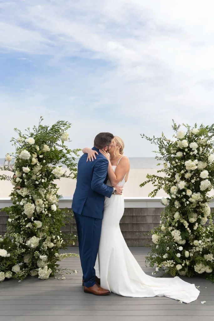 Wychmere Beach Club | Kelsey Sheehan Photography Rhode Island Wedding Photographer | A couple kisses during a seaside wedding, surrounded by lush white floral arrangements. The sky is partly cloudy, adding a serene backdrop to the scene.